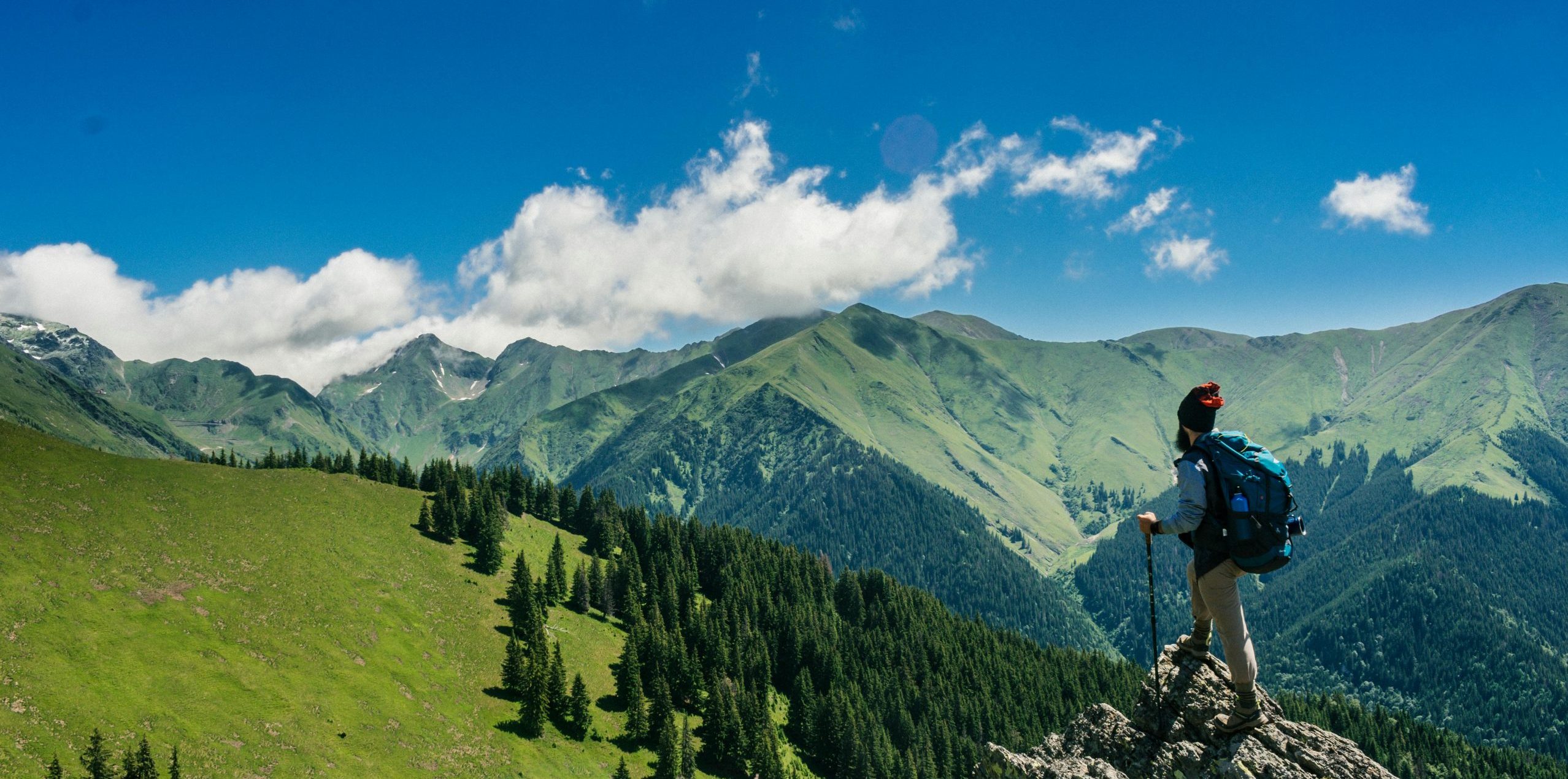 Adventurous hiker enjoying breathtaking views of lush green mountains in Romania under a clear blue sky.